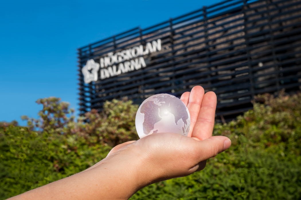 En hand som håller i en glaskula som ser ut som jorden. I bakgrunden gröna buskar och Högskolan Dalarnas fasad.